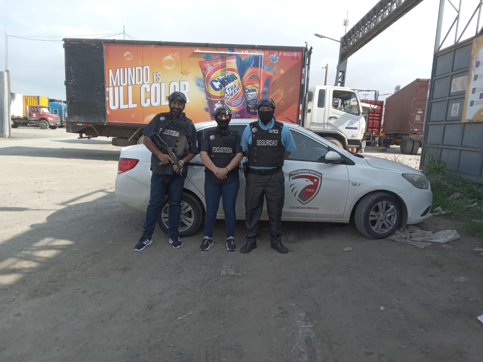 Tres agentes de First Defense con chalecos y fusil junto a un vehículo de custodia entre contenedores
